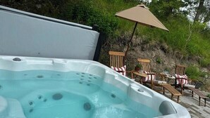Een hottub met een houten terras en stoelen, omringd door begroeiing.