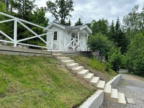 A white wooden cabin with a porch, surrounded by a grassy slope and trees.