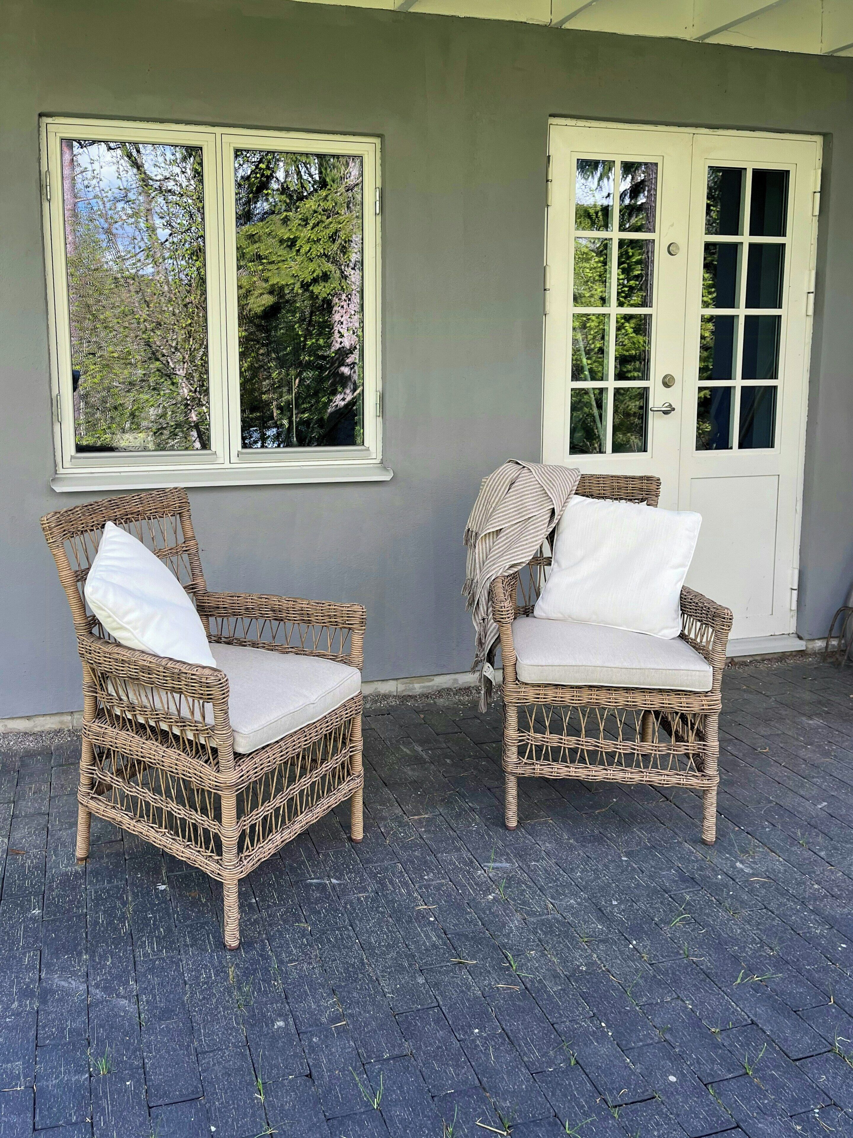 Two wicker chairs with cushions, positioned on a paved patio area with a grey wall and a white door in the background.