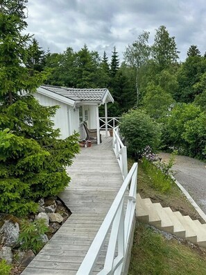 A wooden pathway leading to a white building with a sloped roof, surrounded by greenery.