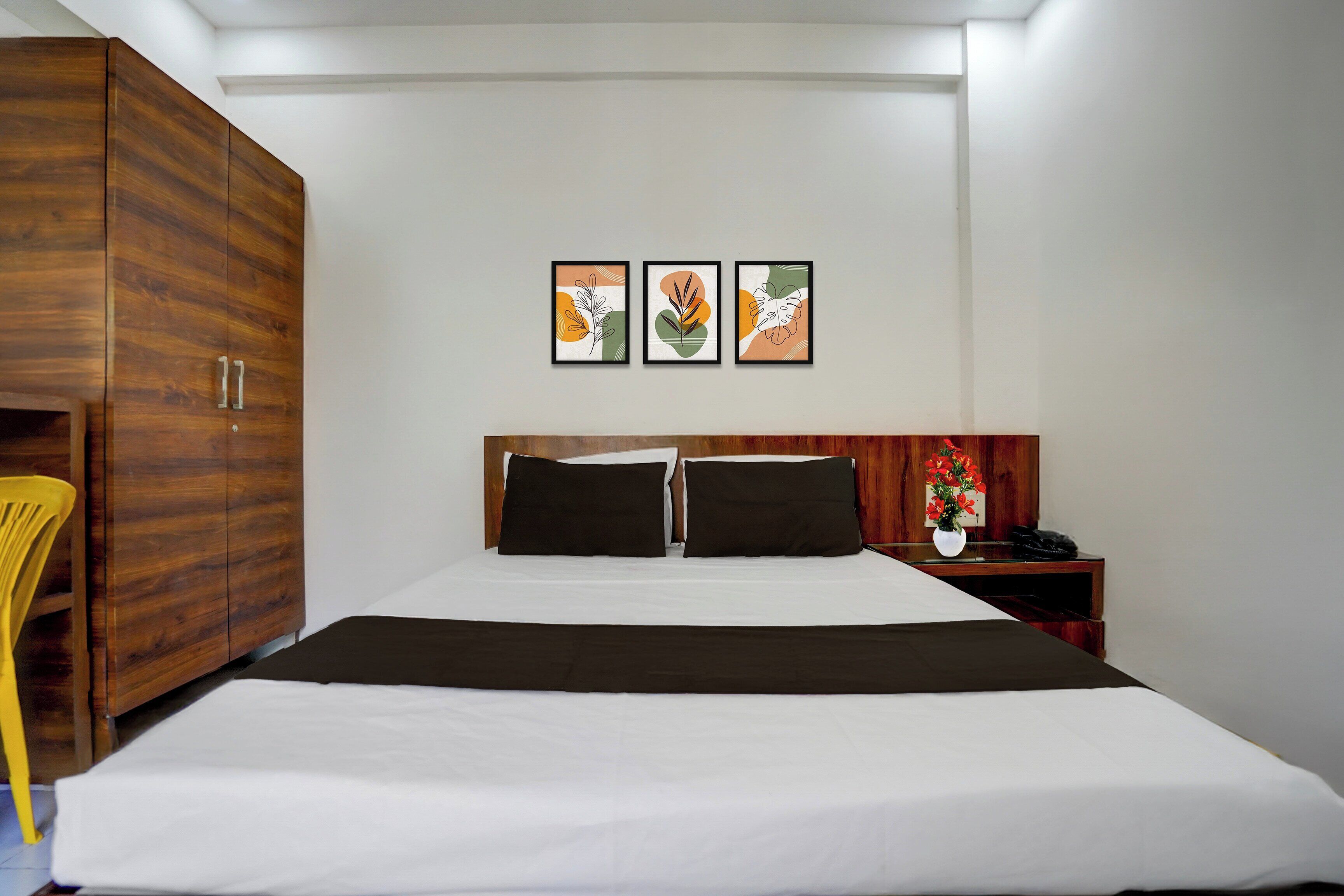 A hotel room with a large bed, a wooden wardrobe, a bedside table with a phone and a vase of flowers, and three framed artworks on the wall.