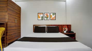 A hotel room with a large bed, a wooden wardrobe, a bedside table with a phone and a vase of flowers, and three framed artworks on the wall.