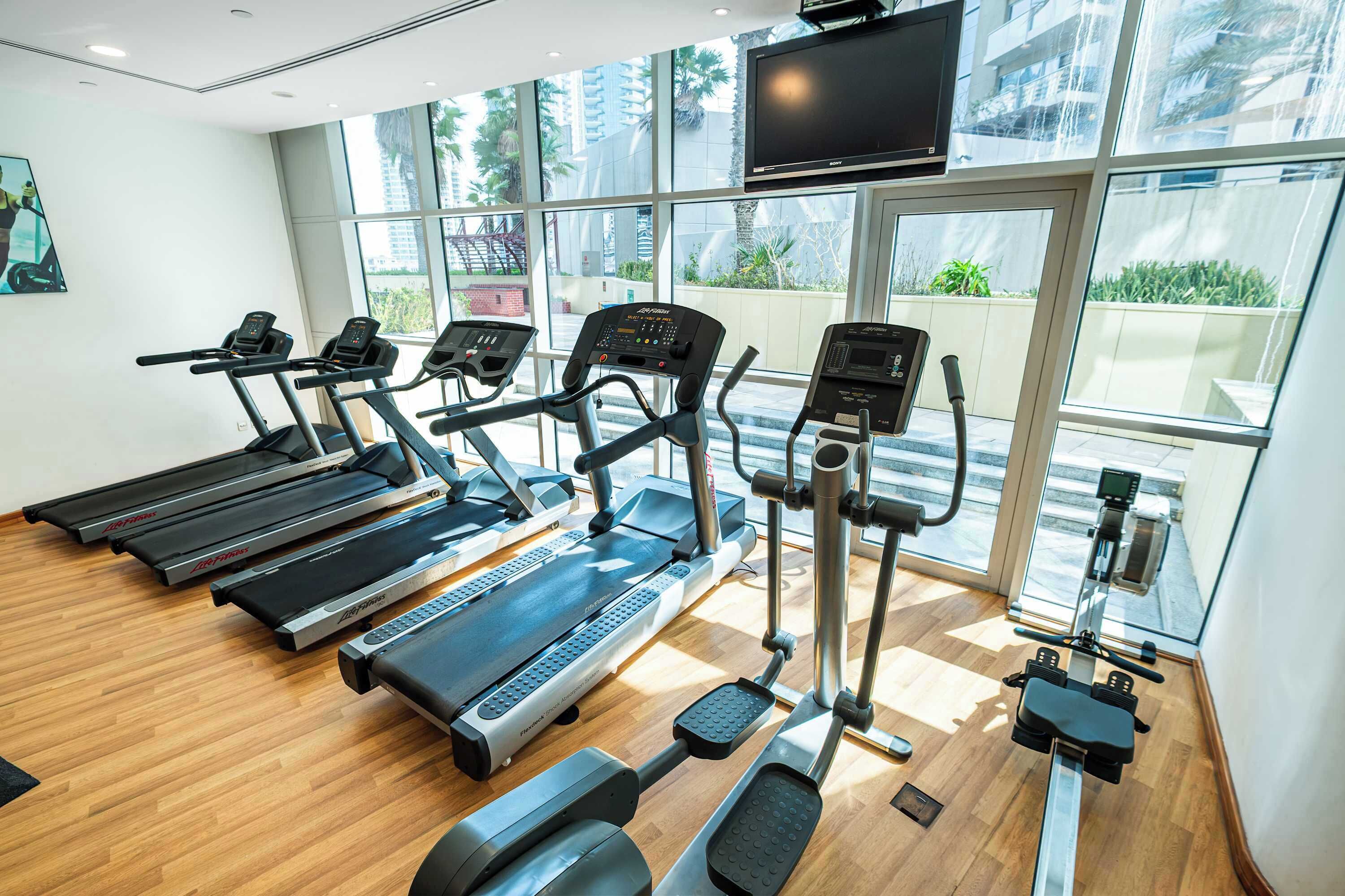 Un gimnasio con cintas de correr, elípticas y una bicicleta estática, con ventanas amplias y un televisor de pantalla plana.