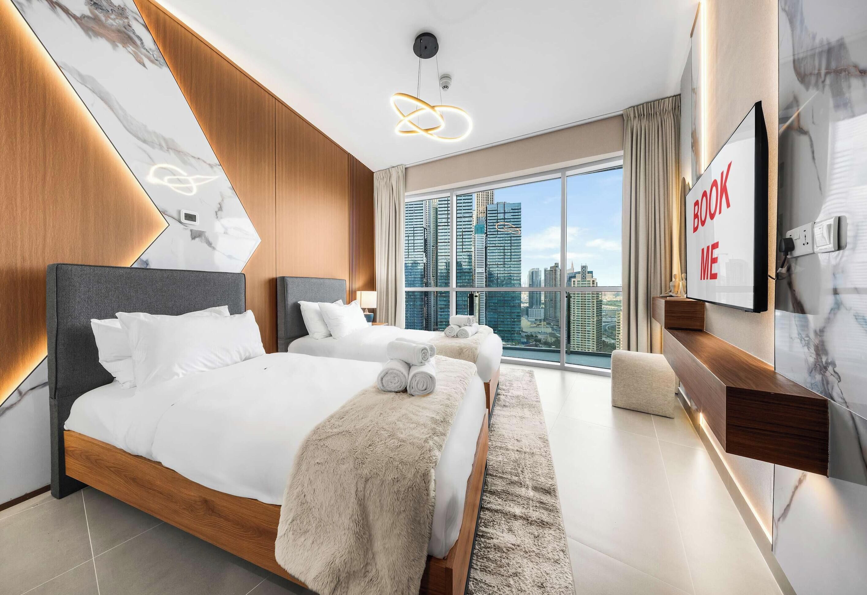 A modern hotel room with a large bed, a smaller bed, a sofa, a TV, and a city view.