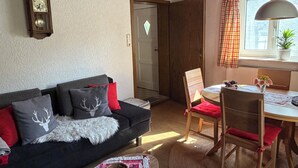 A cozy living room with a dark sofa, a wooden dining table with chairs, a window with curtains, and a wall-mounted clock.