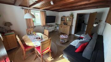 Ein gemütliches Wohnzimmer mit Holzbalkendecke, einem Esstisch mit Stühlen, einer Couch und einem Fernseher.
