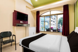 Habitación de hotel con cama, televisor montado en la pared, ventana con cortinas y vista a edificios.