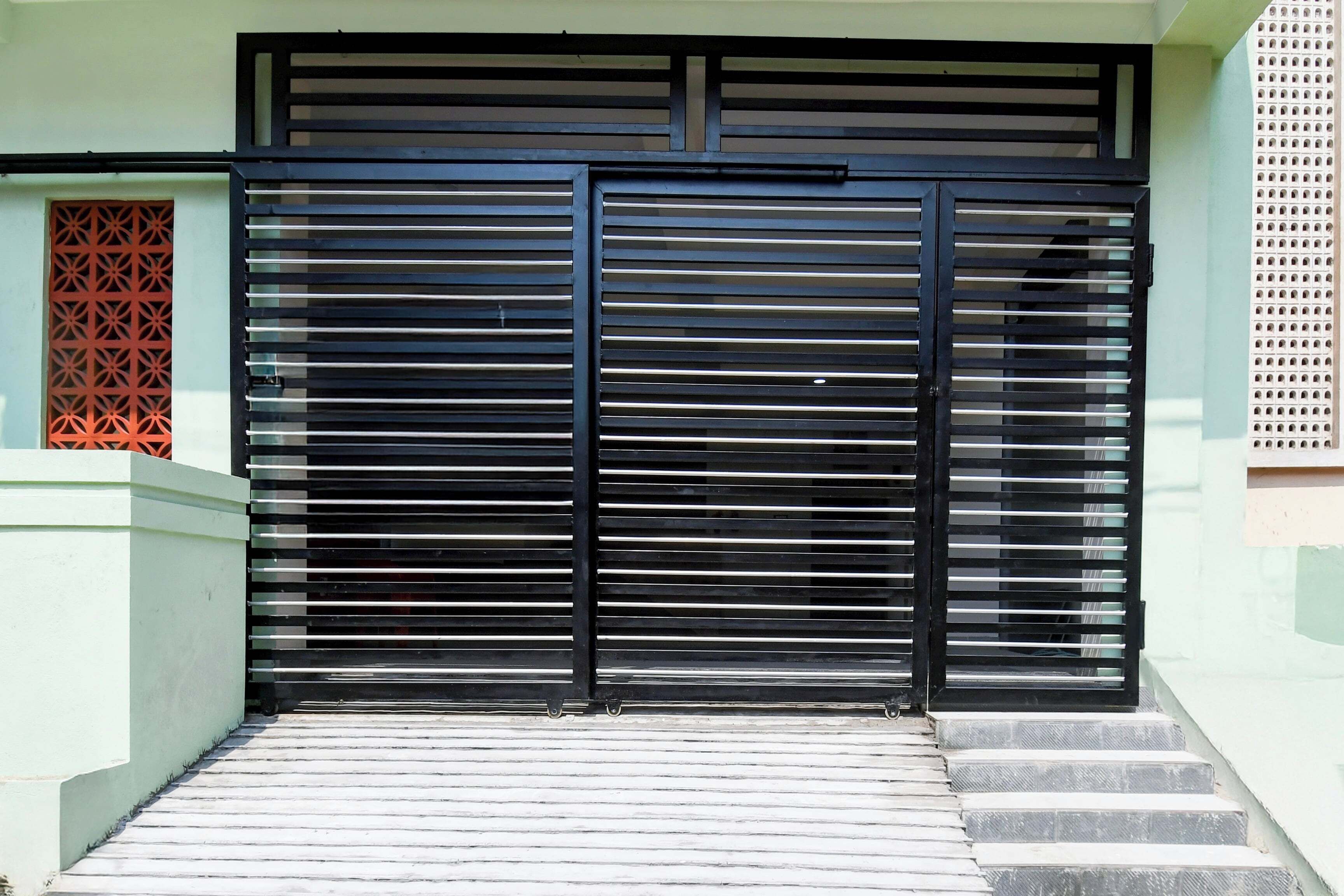Um portão de metal preto com barras horizontais, um lance de escadas que o levam e uma parede branca com padrão geométrico à esquerda.