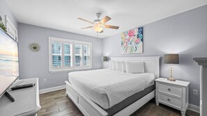 Ein Schlafzimmer mit einem Bett, einem Nachttisch, einer Lampe, einem Deckenventilator, einem Fenster mit Jalousien und einem Bild an der Wand.