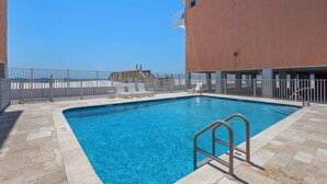 Une piscine avec un escalier en métal, entourée d’une clôture blanche, et un bâtiment avec un balcon en arrière-plan.