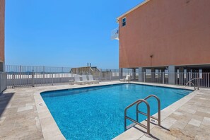 A swimming pool with a metal ladder, surrounded by a white fence, and a building with a balcony in the background. - 2BR Gulf-side condo w private beach access & heated pool (Gulf Shores)