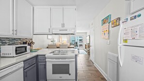 Une cuisine moderne avec des meubles blancs, un four à micro-ondes, une cuisinière et un réfrigérateur. On peut voir divers ustensiles de cuisine sur le plan de travail, et un coin repas est visible à l’arrière-plan.