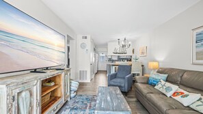 A living room with a television, a sofa, a coffee table, and a kitchen in the background. - 2BR Gulf-side condo w private beach access & heated pool (Gulf Shores)