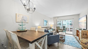 Un salon moderne comprenant un coin repas, un lustre, un tableau représentant un voilier et une vue sur un immeuble à travers une porte vitrée.