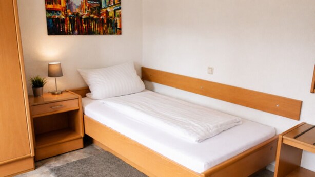 A single bed with a wooden headboard, a bedside table with a lamp, and a small shelf. - Dahlbrucher Hof (Hilchenbach)