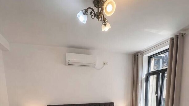 A bedroom with a large bed, bedside tables, a ceiling light fixture, an air conditioning unit, and a window with curtains. - Bright & Elegant 1BR Apartment | City Center (Bucharest)