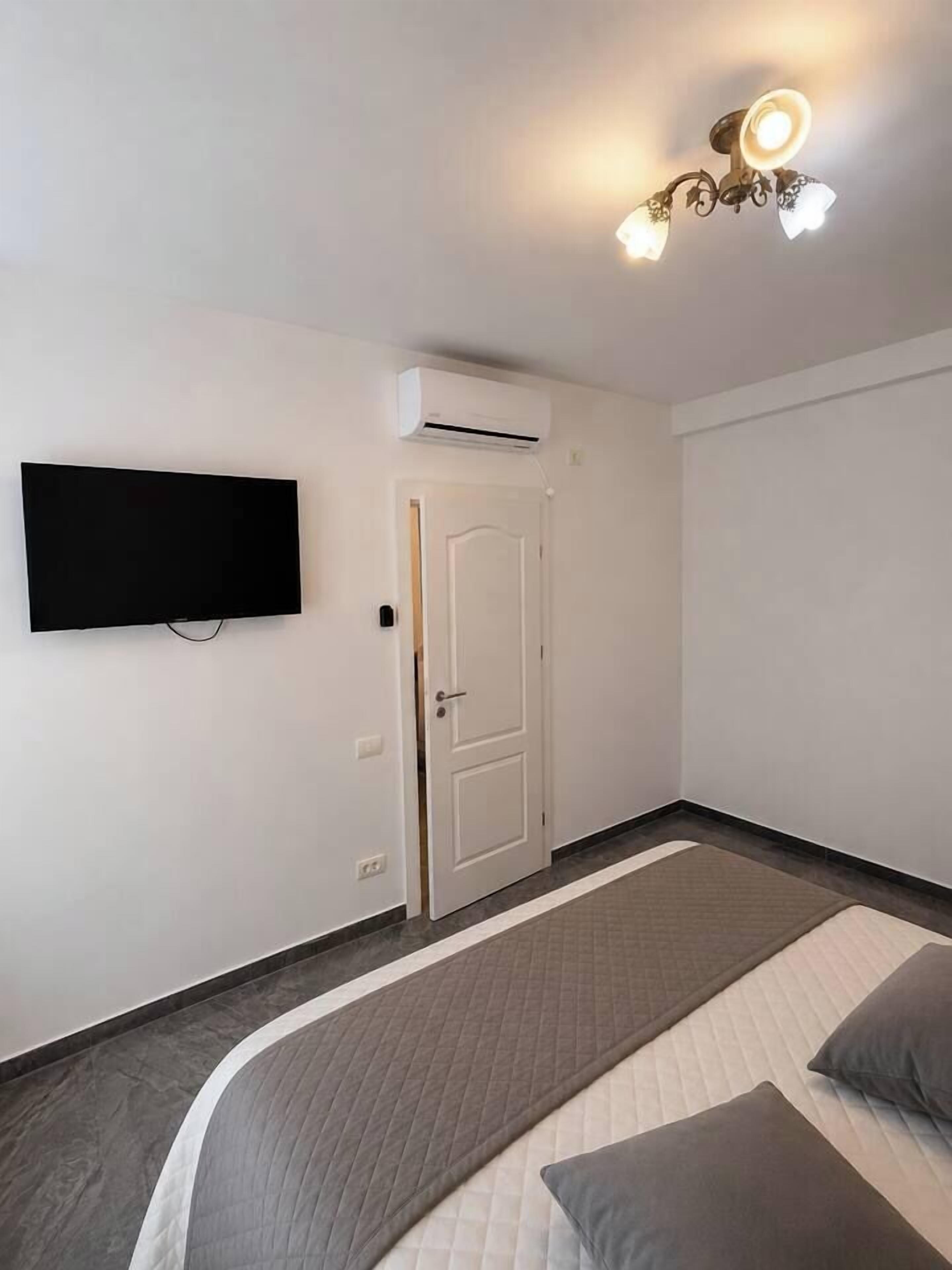 A bedroom with a bed, a flat-screen TV, an air conditioning unit, a door, and a ceiling light fixture.