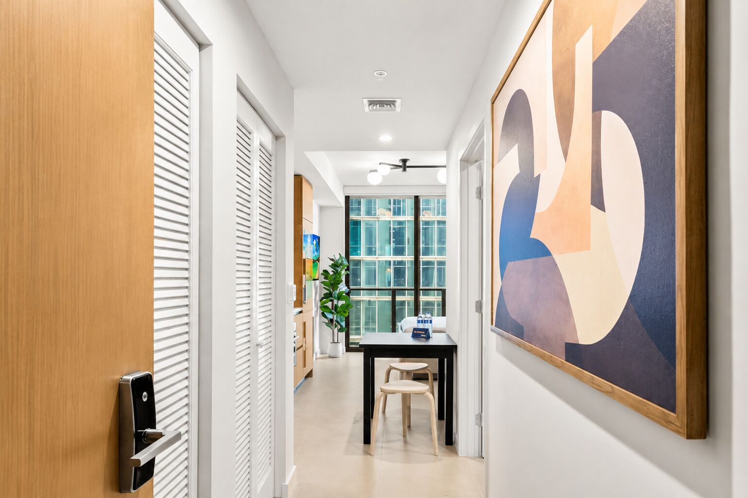 Un pasillo de hotel con una puerta de madera, una pared blanca con un cuadro abstracto grande, una planta en maceta y un escritorio con una silla.
