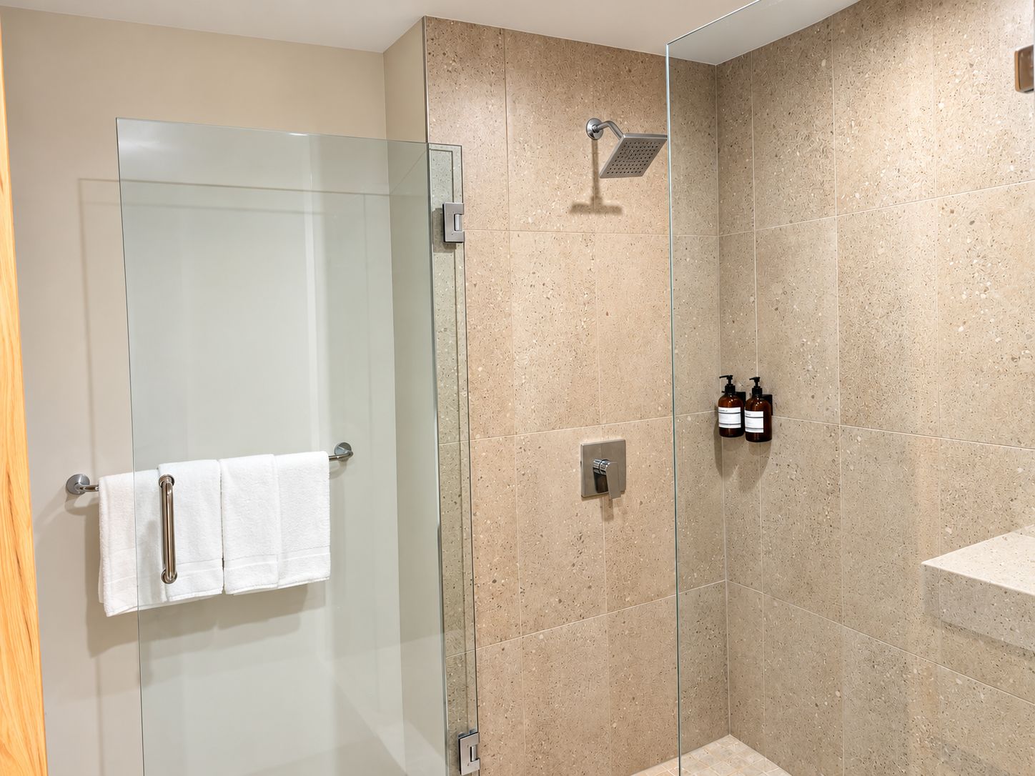 Une douche fermée par des parois en verre, dotée d’un pommeau de douche à main, d’un porte-serviettes avec deux serviettes et de deux bouteilles de shampoing fixées au mur.