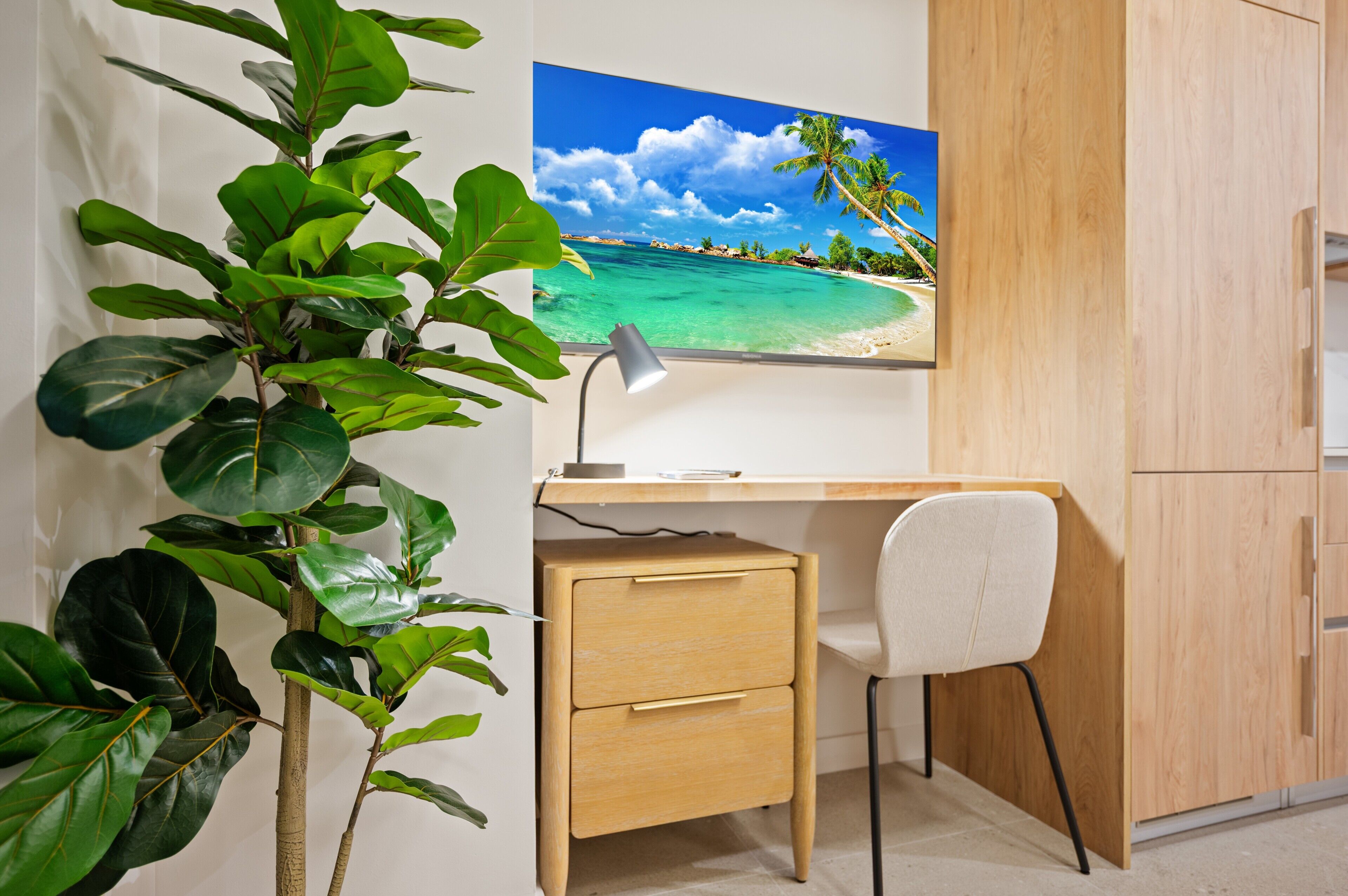 A room with a desk, a chair, a drawer, a plant, and a wall-mounted TV displaying a beach scene.