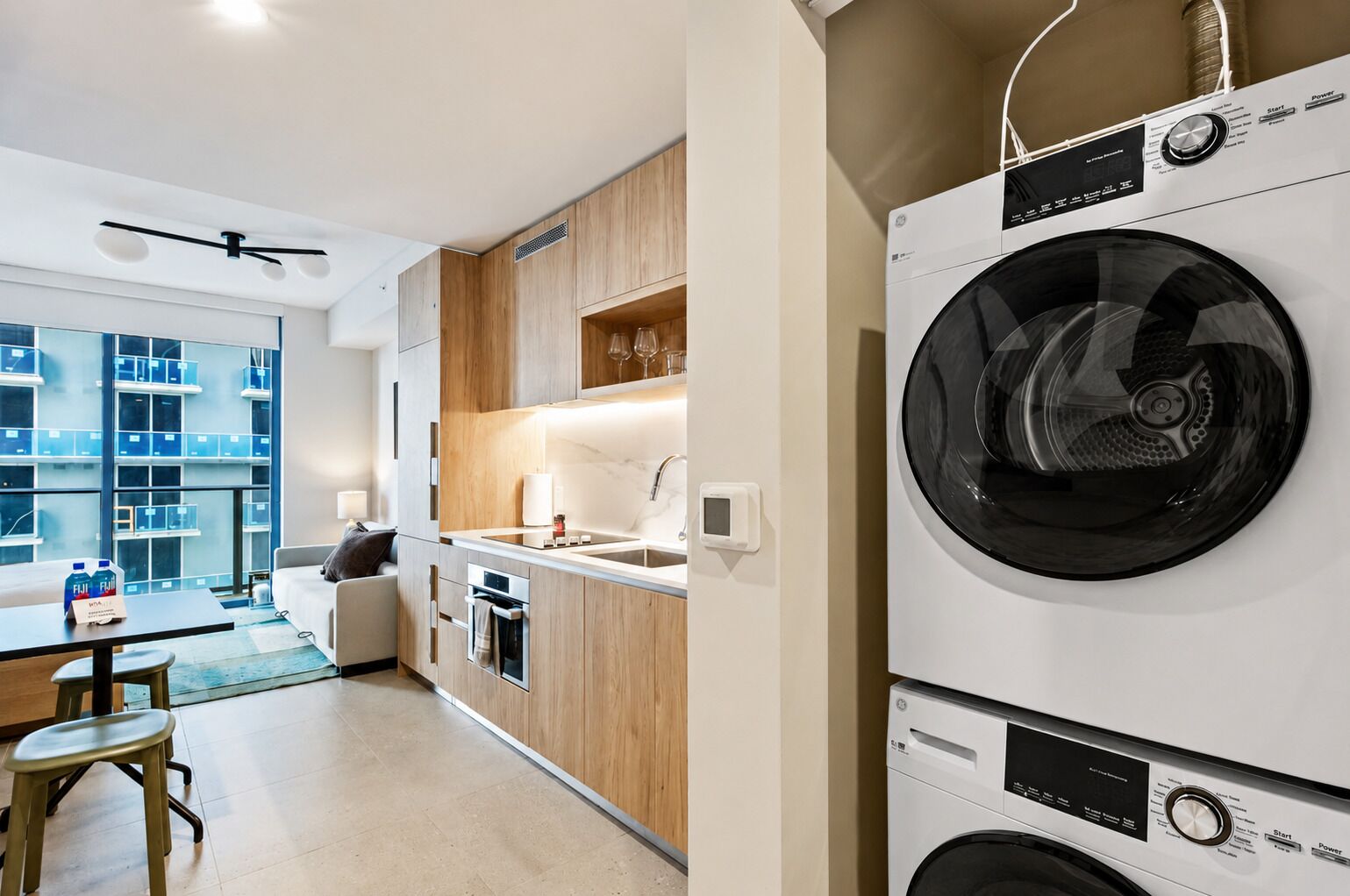 Een moderne keuken met een ingebouwde wasmachine en droger, een eethoek met tafel en stoelen, en uitzicht op een woongebouw door grote ramen.
