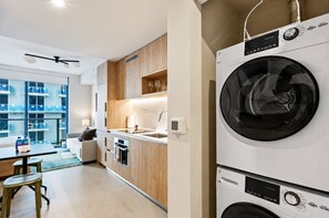 Une cuisine moderne équipée d’un lave-linge et d’un sèche-linge intégrés, un coin repas avec une table et des chaises, et une vue sur un immeuble d’habitation à travers de grandes fenêtres.