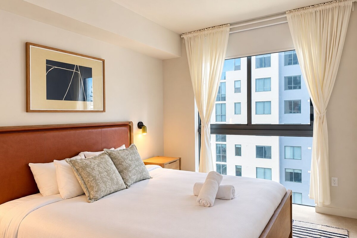 Une chambre d’hôtel avec un grand lit, un tableau encadré au mur, une table de chevet avec une lampe et une vue sur un immeuble par la fenêtre.