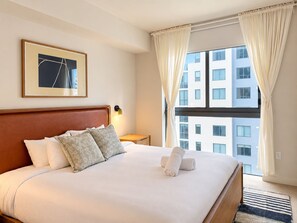 Une chambre d’hôtel avec un grand lit, un tableau encadré au mur, une table de chevet avec une lampe et une vue sur un immeuble par la fenêtre.