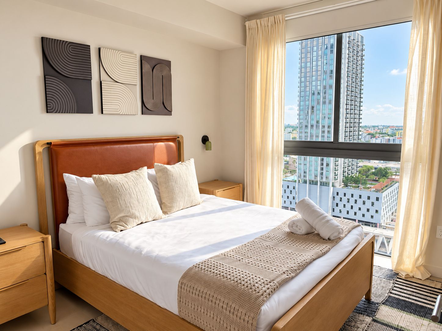 Habitación de hotel con una cama grande, una mesita de noche, una lámpara y vistas de edificios altos.