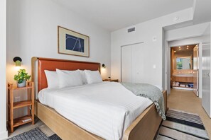 Una cama bien tendida con sábanas blancas, un cabecero de madera y una mesita de noche con una lámpara y una pequeña planta.