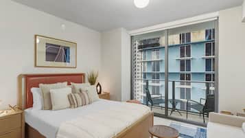 Un dormitorio moderno con una cama grande, una mesita de noche, una silla, una pequeña mesa redonda y un balcón con vistas a un edificio.