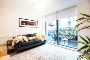 Uma sala de estar moderna com um sofá, um quadro emoldurado, um abajur e uma janela grande com vista para um edifício e áreas verdes.