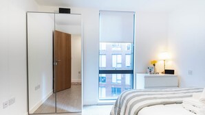 Quarto com cama, mesa de cabeceira com abajur, espelho e janela com vista para edifícios.