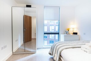 Ein Schlafzimmer mit einem Bett, einem Nachttisch mit Lampe, einem Spiegel und einem Fenster mit Blick auf Gebäude.