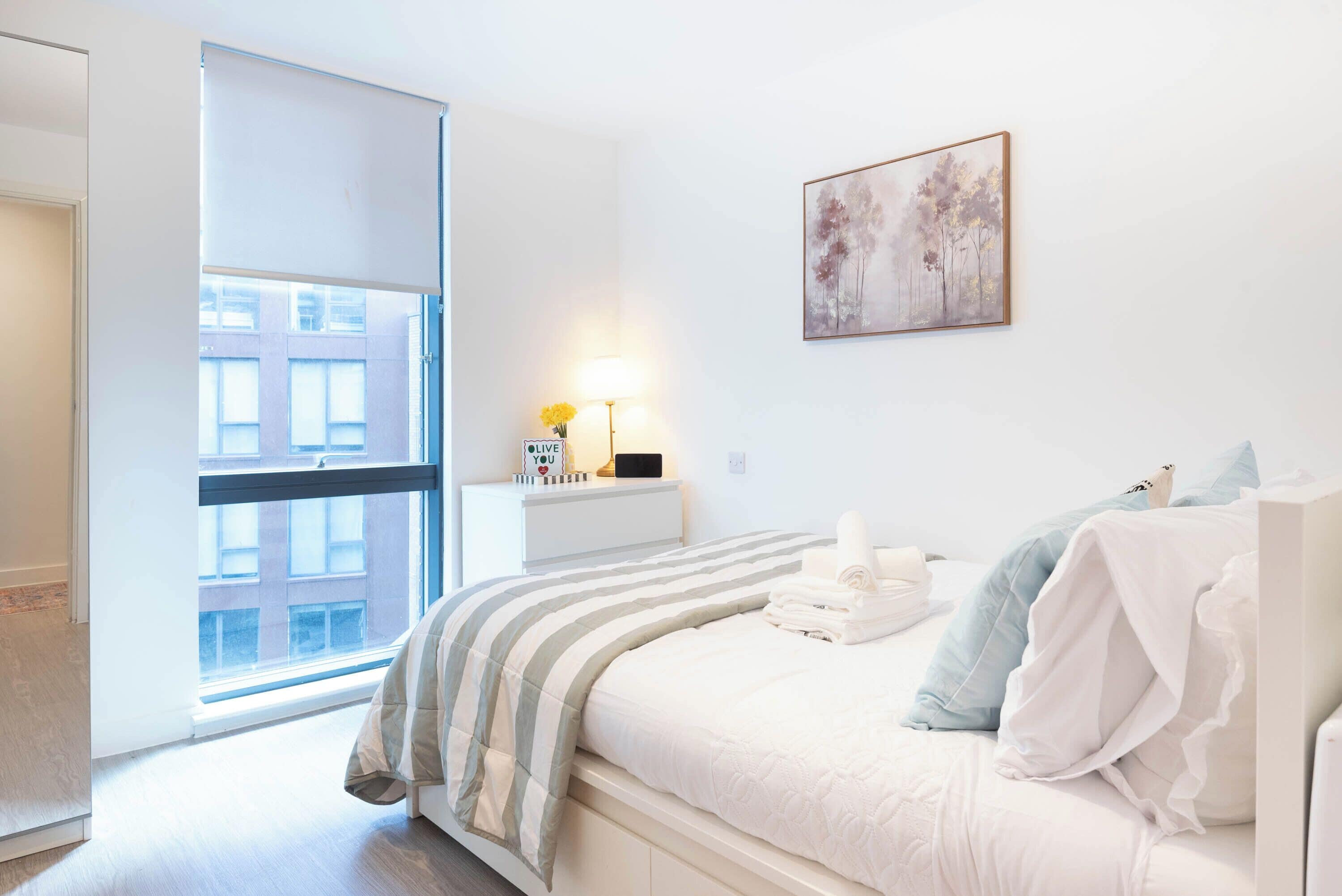 A hotel room with a bed, a bedside table with a lamp, a painting on the wall, and a view of buildings outside the window.