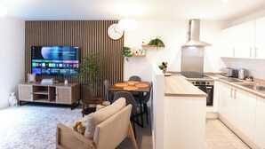 Uma sala de estar moderna com televisão de tela plana, uma área de jantar com mesa de madeira e uma cozinha com armários brancos e coifa de aço inoxidável.