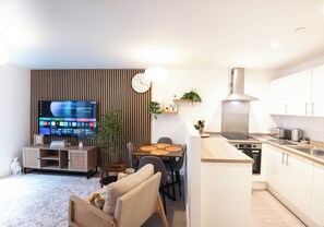 Ein modernes Wohnzimmer mit Flachbildfernseher, einem Essbereich mit Holztisch und einer Küche mit weißen Schränken und einer Edelstahl-Dunstabzugshaube.