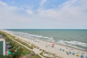 A beach with umbrellas and people, a road parallel to the beach, and a building on the left. - Oceanfront 1BR Suite + Beach Access + Lazy River (Myrtle Beach)