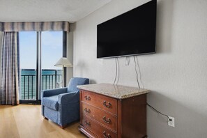A room with a flat-screen TV mounted on the wall, a blue armchair, a wooden dresser with drawers, and a view of the sea through a balcony with curtains. - Oceanfront 1BR Suite + Beach Access + Lazy River (Myrtle Beach)