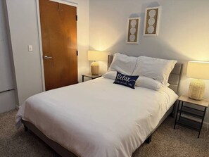 Een netjes opgemaakt bed met een kussen waarop staat 'Ontspan, je bent op vakantie aan het meer' in een hotelkamer met een houten deur, twee aan de muur bevestigde fotolijstjes en twee bedlampjes.
