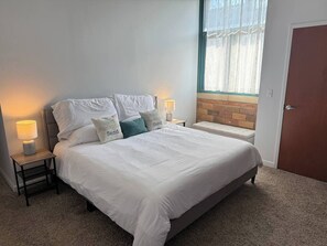 A neatly made bed with white bedding and multiple pillows, two bedside lamps, a small nightstand, and a window with a view of a brick wall. - Spacious two bedroom with pool (Sandusky)