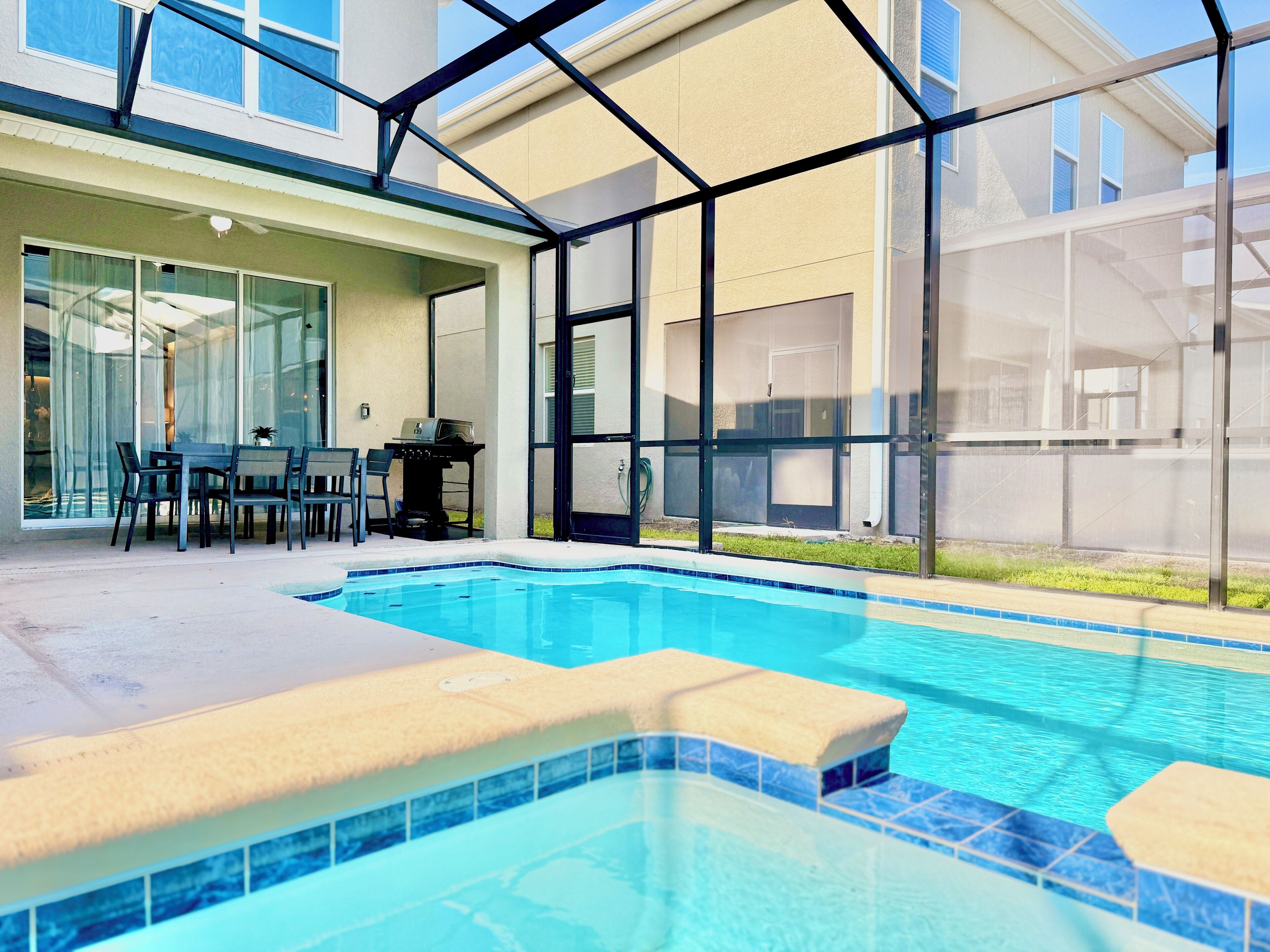 A swimming pool with a tiled edge, a patio area with a table and chairs, and a covered outdoor space with a grill.