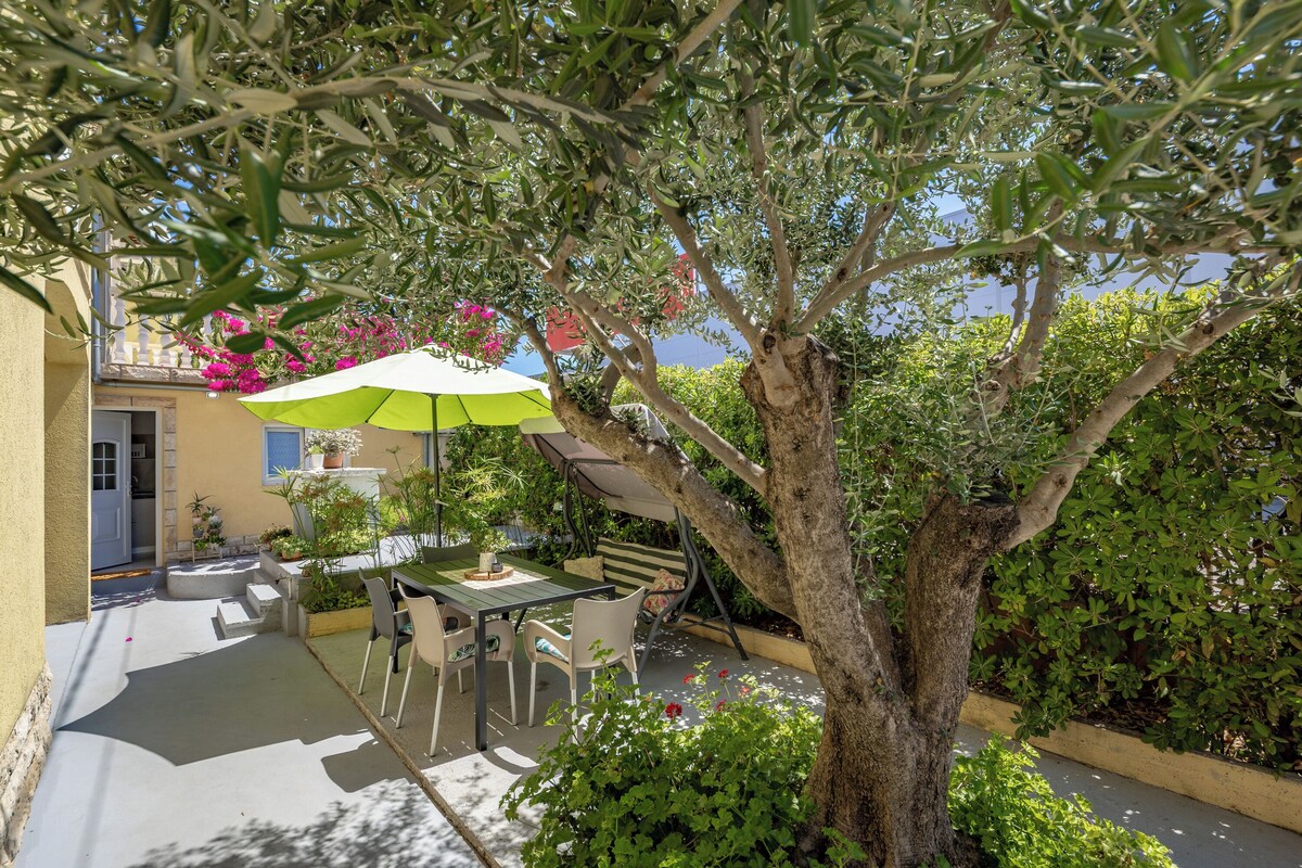 An outdoor patio with a table and chairs, surrounded by greenery and a tree.