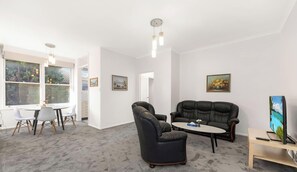 Una sala de estar con un sofá de cuero negro, una mesa redonda y un televisor sobre un mueble bajo. Hay una zona de comedor con una mesa y sillas blancas.