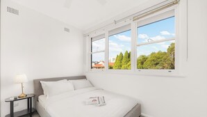A hotel room with a bed, a nightstand, a lamp, and a window with a view of trees and buildings. - Metawise 2BR Apt Brightest in Brighton (Sydney)