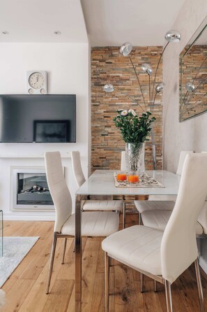 A modern dining area with a white table, four chairs, a vase with flowers, and a flat-screen TV above a fireplace. - Modern 3-bedroom Home Near Westfield Central Lond (London)