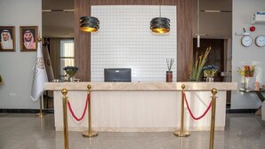 Un comptoir d’accueil surmonté d’un plateau en marbre, flanqué de deux stanchions dorées reliées par des cordes rouges, avec un drapeau et une horloge murale.