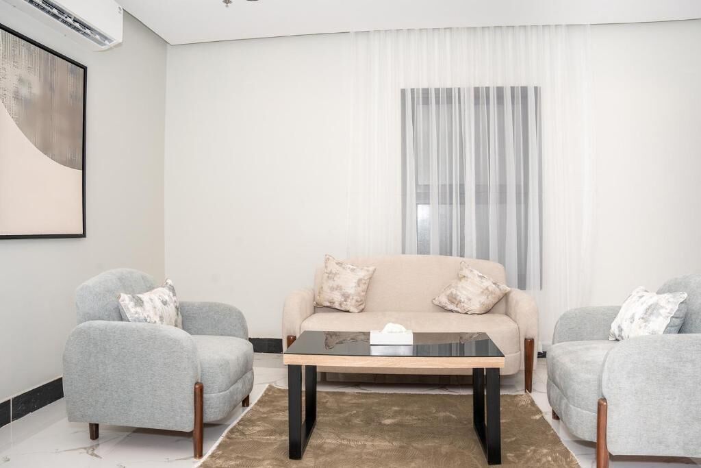 Un salón con un sofá color crema, dos sillones grises, una mesa de centro de vidrio y un ventanal con cortinas de gasa.