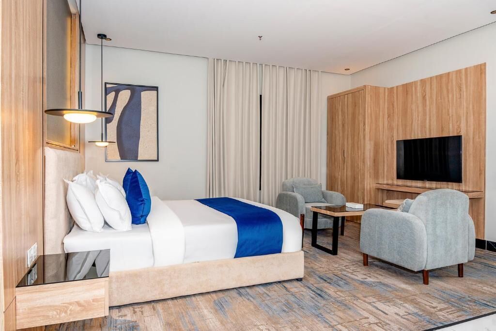 Habitación de hotel con cama, zona de estar con mesa, televisor y un amplio ventanal con cortinas.