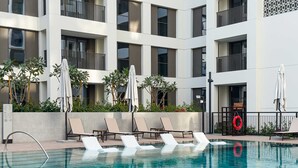 A swimming pool with lounge chairs and umbrellas, surrounded by a building with balconies and greenery. - Free Housekeeping Visits During Your Stay - Stayshort - Peaceful 1-bedroom in Dubai Creek Harbour - Sleeps 4 (Dubai)